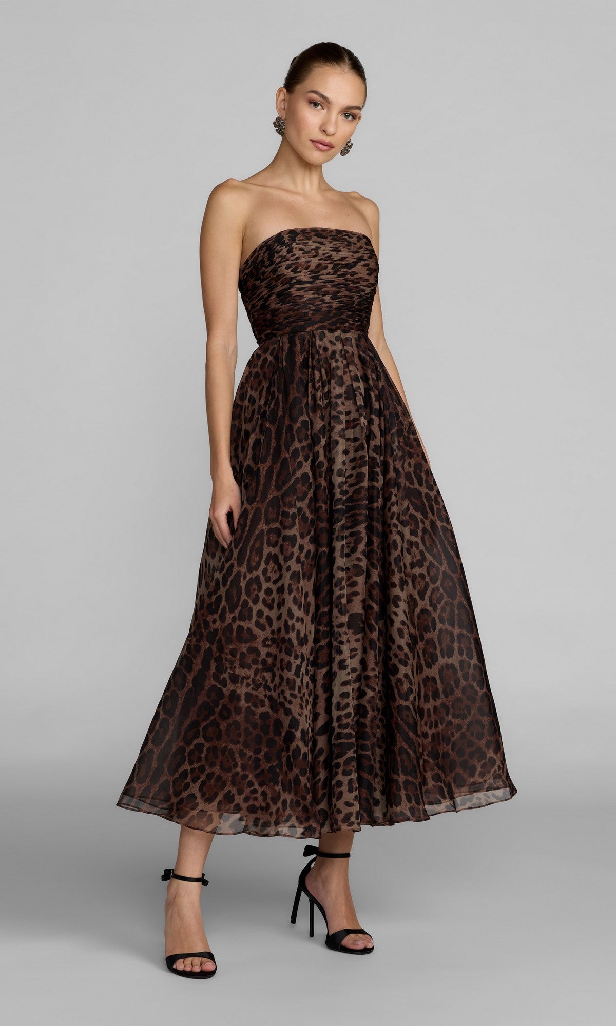 A woman stands against a light gray background wearing the Strapless Leopard Formal Dress: Mac Duggal 56350—an ankle-length, leopard print design with an a-line skirt—paired with black high-heeled sandals.